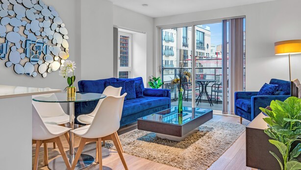 A modern living room with a glass coffee table, blue sofas, and a dining area with white chairs. - MySquare Apartments Shoredtich (London)