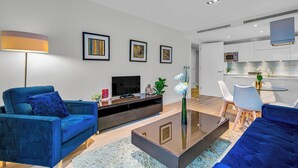 A modern living room with a blue sofa, a coffee table, and a TV. There is a kitchen area with white cabinets and a microwave. - MySquare Apartments Shoredtich (London)