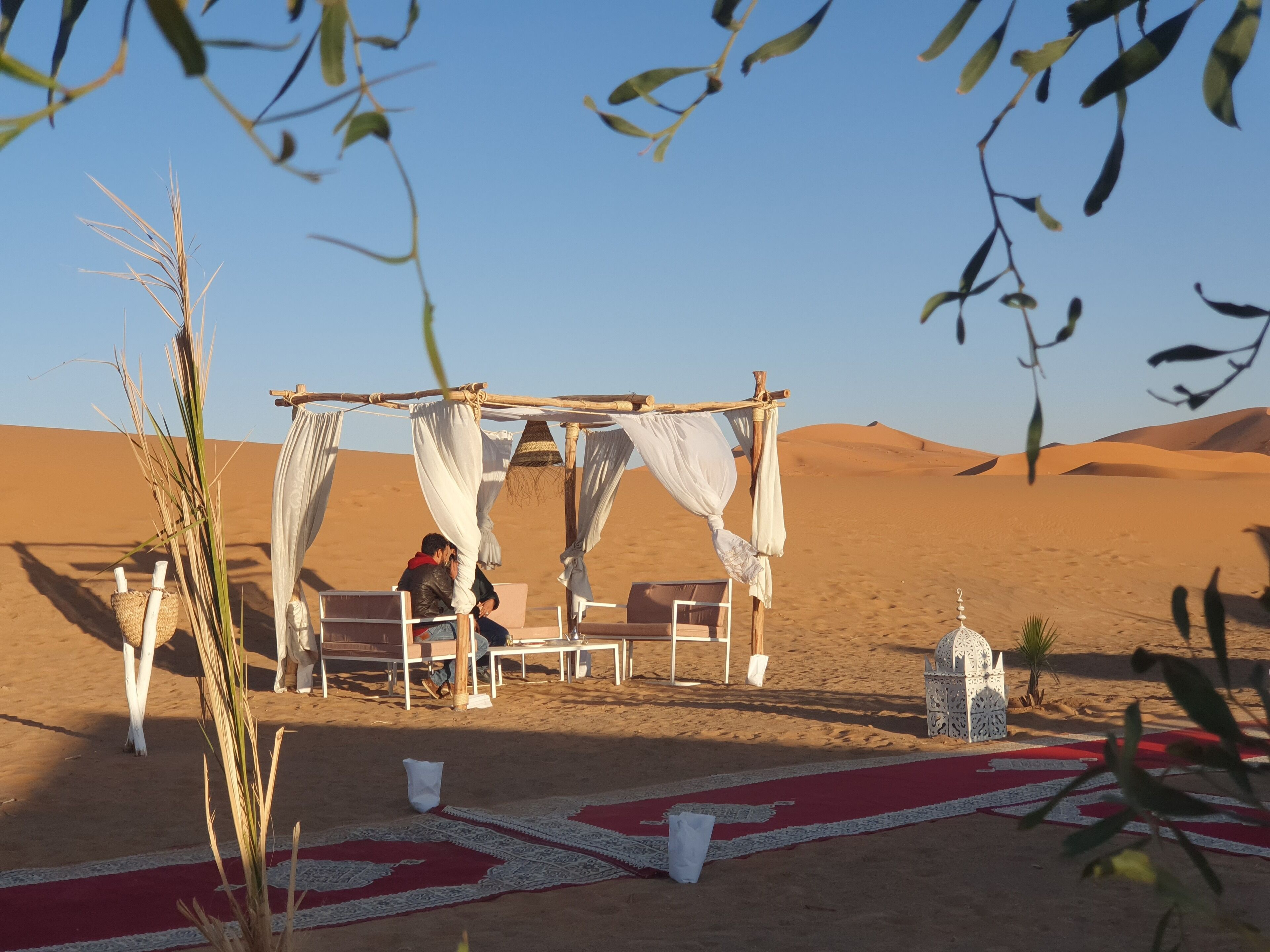 Una tienda de campaña en el desierto con alfombra roja y cortinas blancas, frente a dunas de arena.