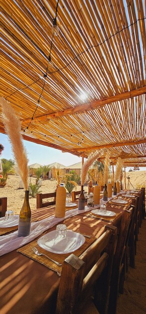 An outdoor dining area with bamboo ceiling and tables set for a meal, overlooking a desert landscape. - Tangerine Sky Camp (Rissani)