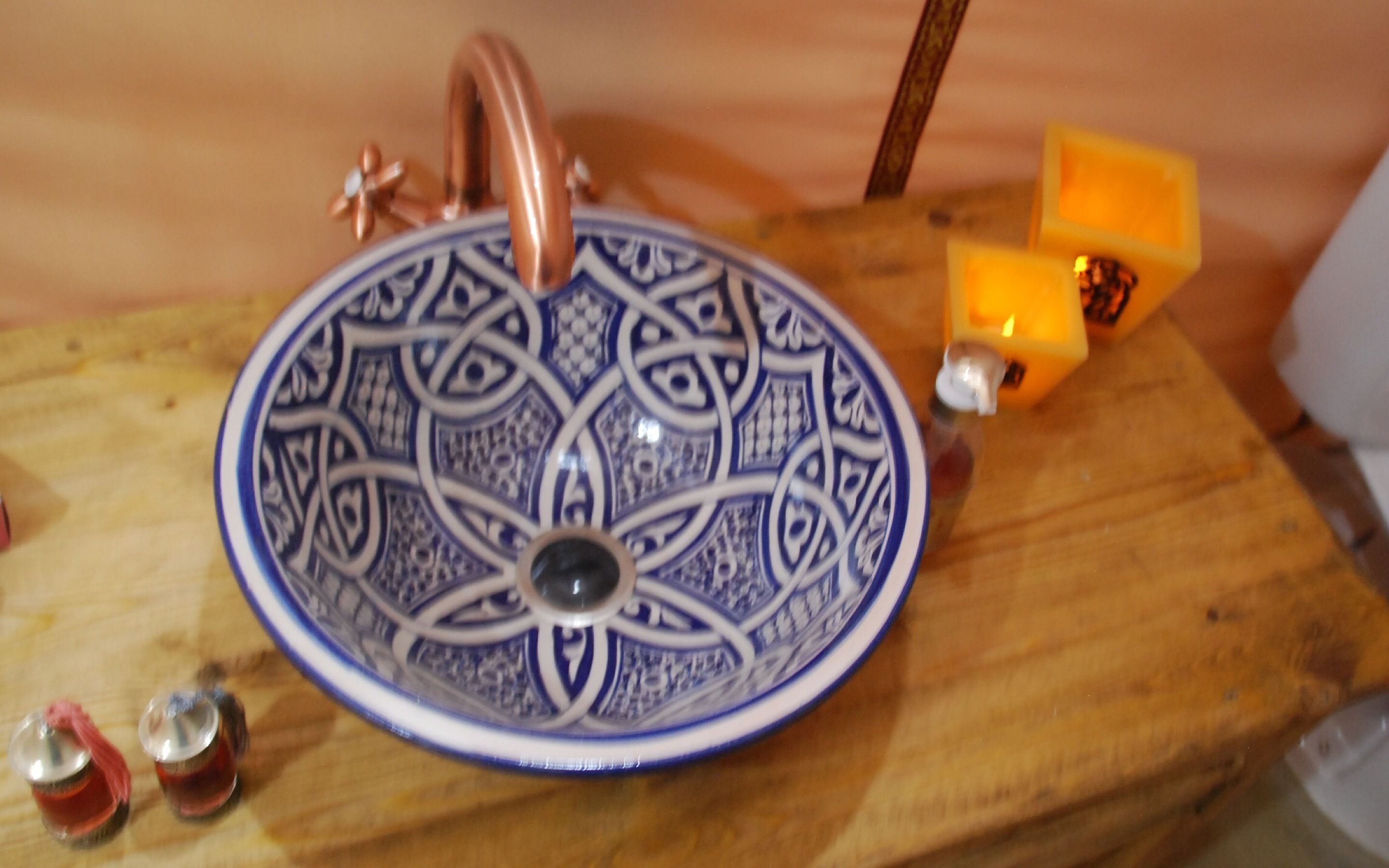 Uma pia de cerâmica com padrões azuis elaborados, uma torneira de cobre e um castiçal laranja pequeno sobre uma superfície de madeira.