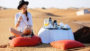 Una persona sentada en un cojín rojo en un paisaje desértico, con una mesa dispuesta para una comida.