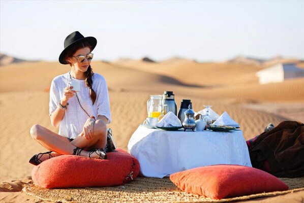 A person sitting on a red cushion in a desert setting with a table set for a meal. - Tangerine Sky Camp (Rissani)