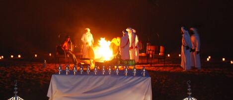 A desert wedding setup with a table covered in white cloth, surrounded by guests and a central fire.