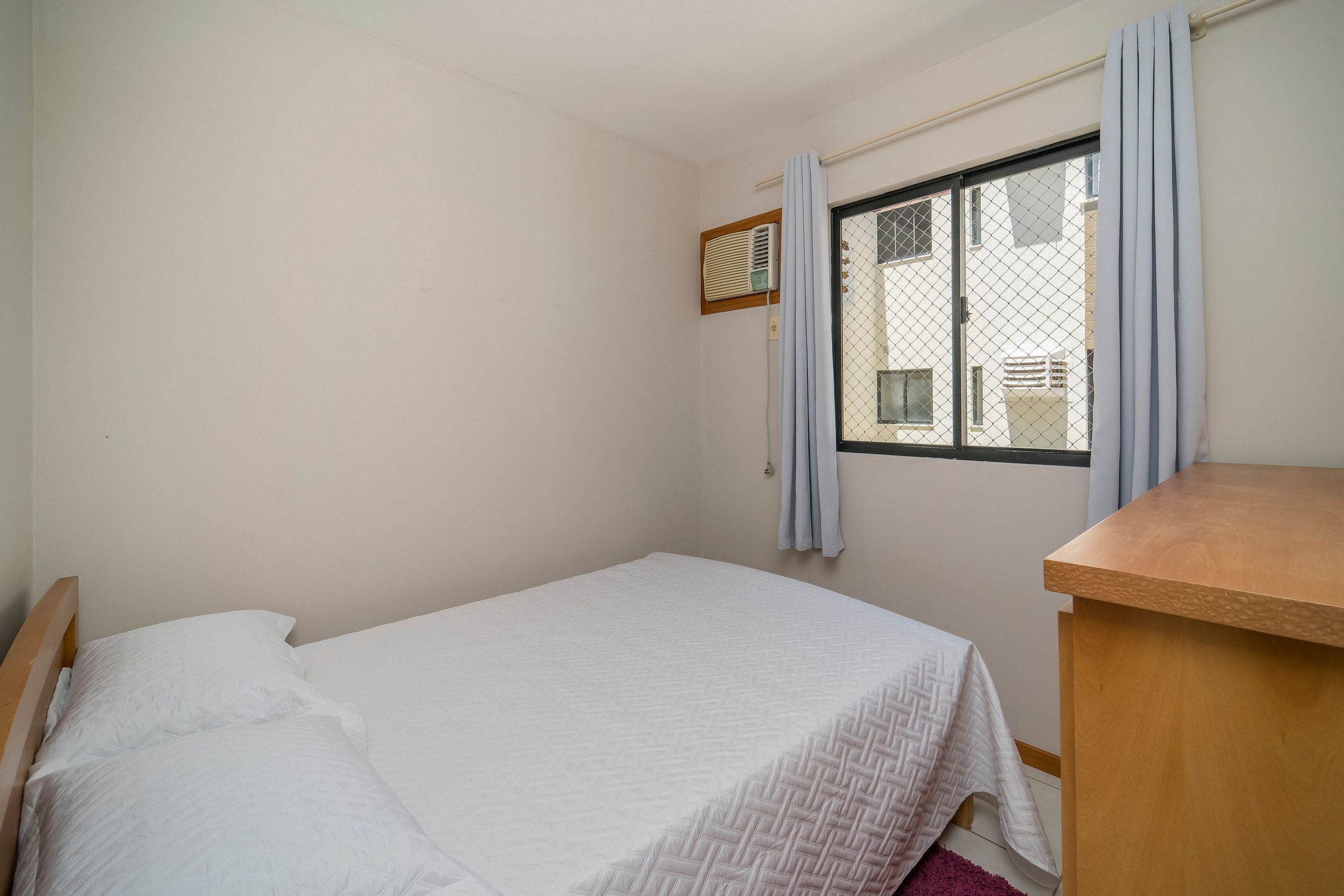 A small, clean room with a single bed, a wooden nightstand, a window with curtains, and a wall-mounted air conditioning unit.