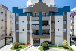 A multi-story residential building with balconies, a central entrance, and a small garden area.