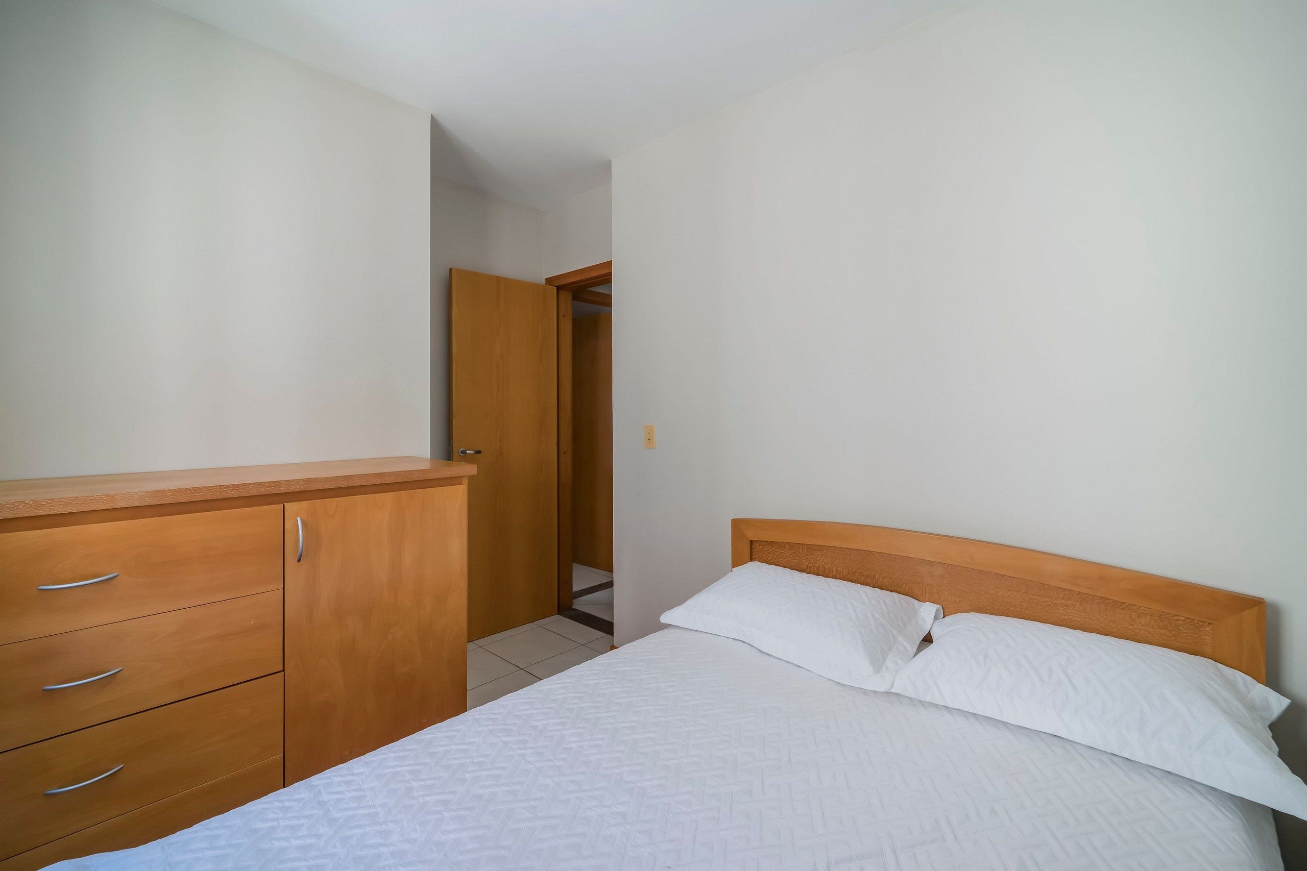 Un dormitorio con cabecera de cama de madera, ropa de cama blanca y un aparador de madera con cajones.