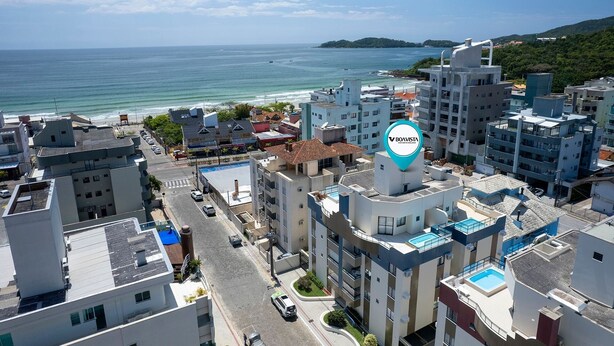 A coastal urban area with modern buildings, a clear view of the sea, and a prominent BOAVISTA logo on a building. - Apartamento 2 quartos 1 suíte - 081 (Bombinhas)