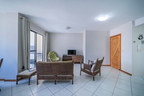 A living room with wicker furniture, a television, and a wooden door. - Apartamento 2 quartos 1 suíte - 081 (Bombinhas)