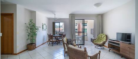 A spacious living area with a dining table, wicker chairs, a green armchair, a TV, and a potted plant.