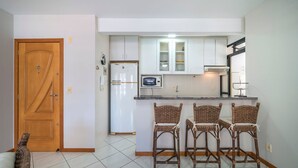 A kitchen with white cabinets, a refrigerator, a microwave, and three wicker bar stools. - Apartamento 2 quartos 1 suíte - 081 (Bombinhas)