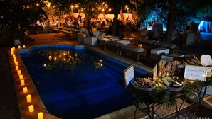 A poolside area with seating and tables, surrounded by trees and illuminated by string lights.