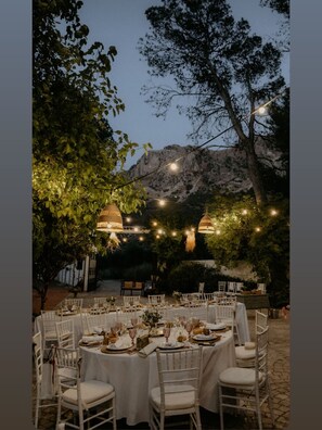 Un espacio exterior para comer con mesas y sillas redondas, luces colgantes y un paisaje montañoso de fondo.