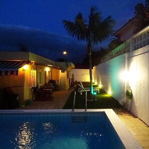 Una piscina con una palmera, un porche techado y una pared blanca con iluminación decorativa.