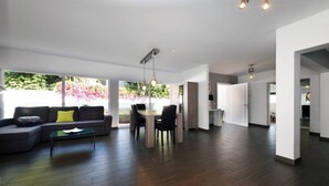 A spacious living area with a dark wood floor, a sectional sofa with a yellow pillow, a glass coffee table, and a dining area with a table and chairs. There is a large window providing a view of greenery outside. - Taoro House, Puerto de la Cruz (Puerto de la Cruz)