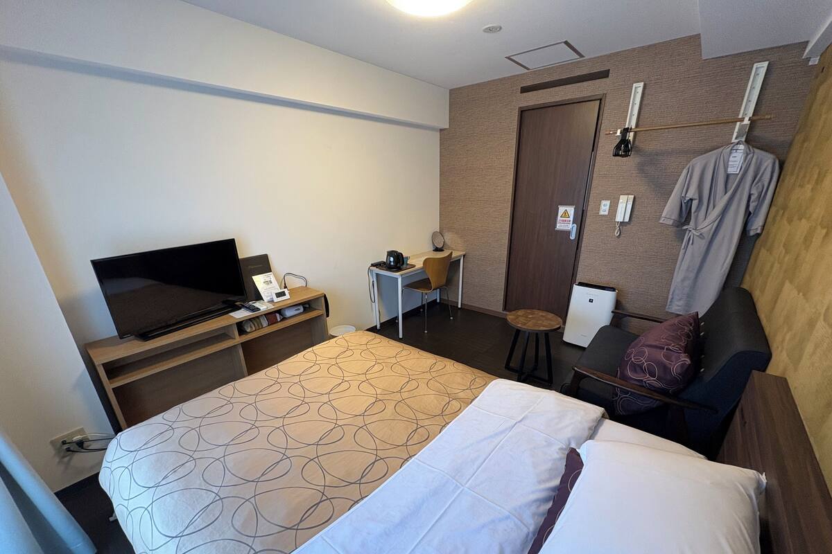 A hotel room with a bed, a desk with a television, a small table, a chair, and a wardrobe.