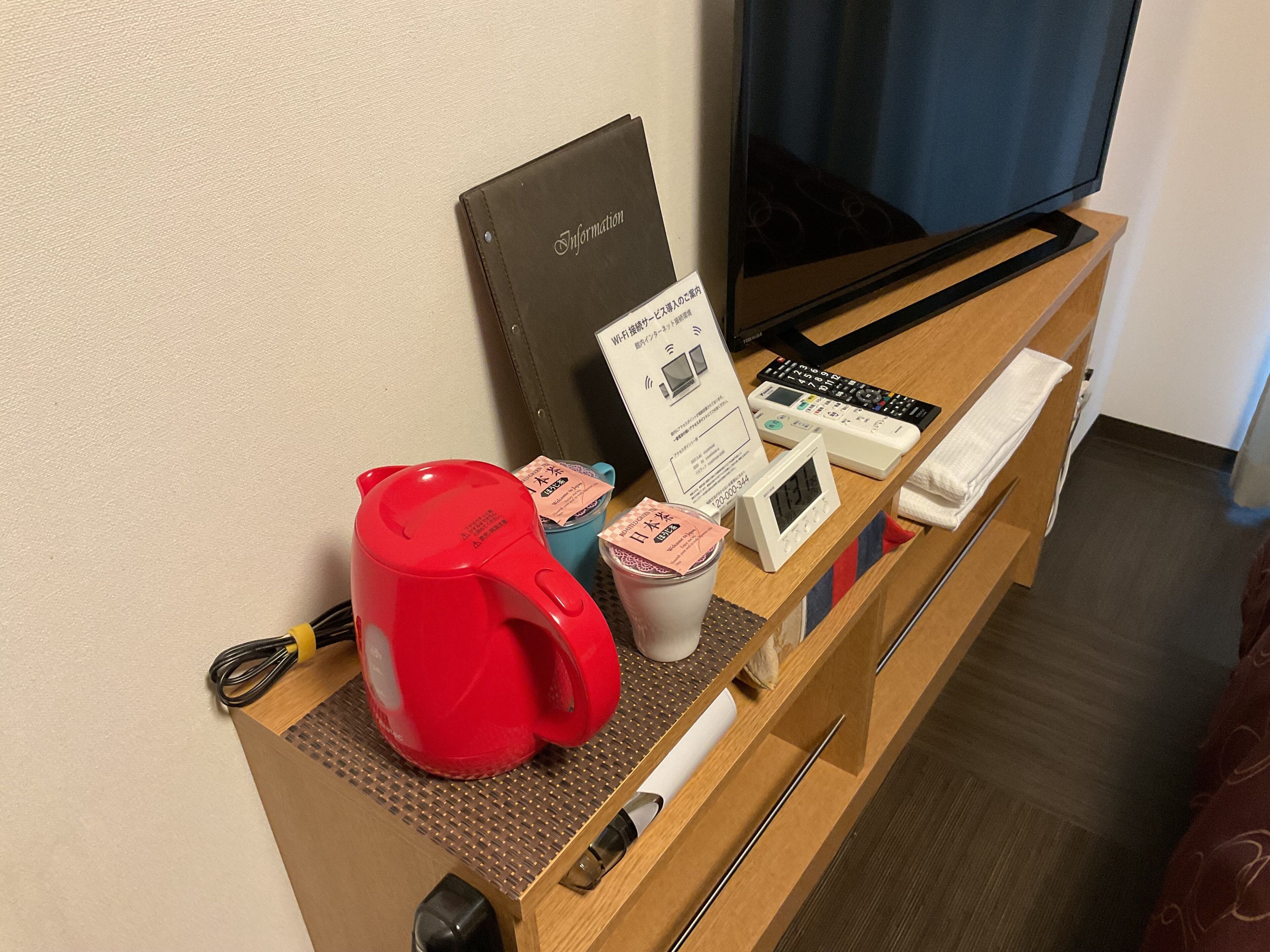 A hotel room with a wooden desk, a red kettle, a digital clock, a remote control, and a television.
