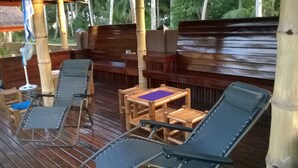 Een overdekt terras met twee loungestoelen en een kleine tafel, omgeven door houten leuningen en een bamboestructuur.