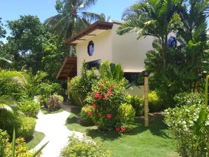 A house with a white exterior, a brown roof, and a small window. It is surrounded by lush greenery, including palm trees and various plants. - Sunset Bay Villas Siargao (General Luna)