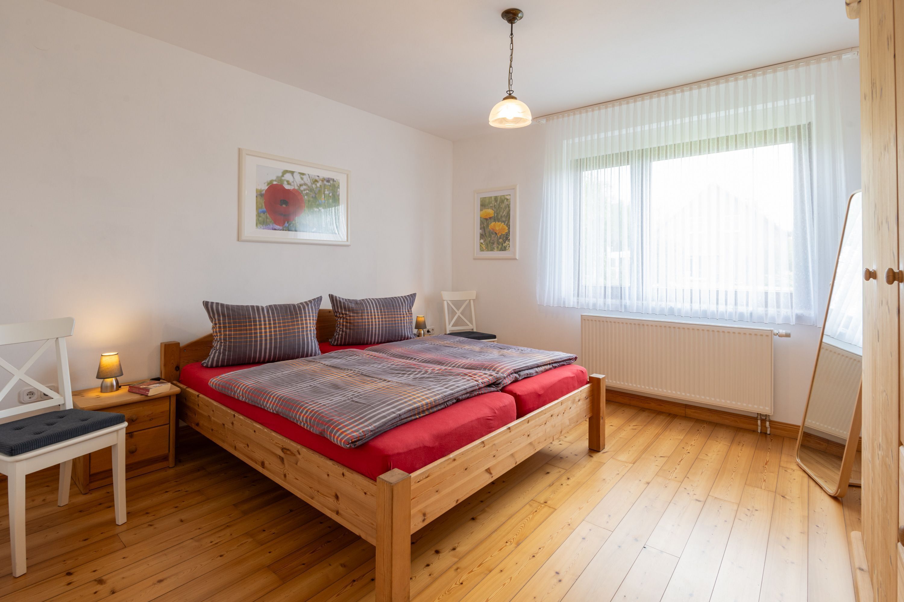 A bedroom with a wooden bed, a nightstand, a chair, and a window with sheer curtains.