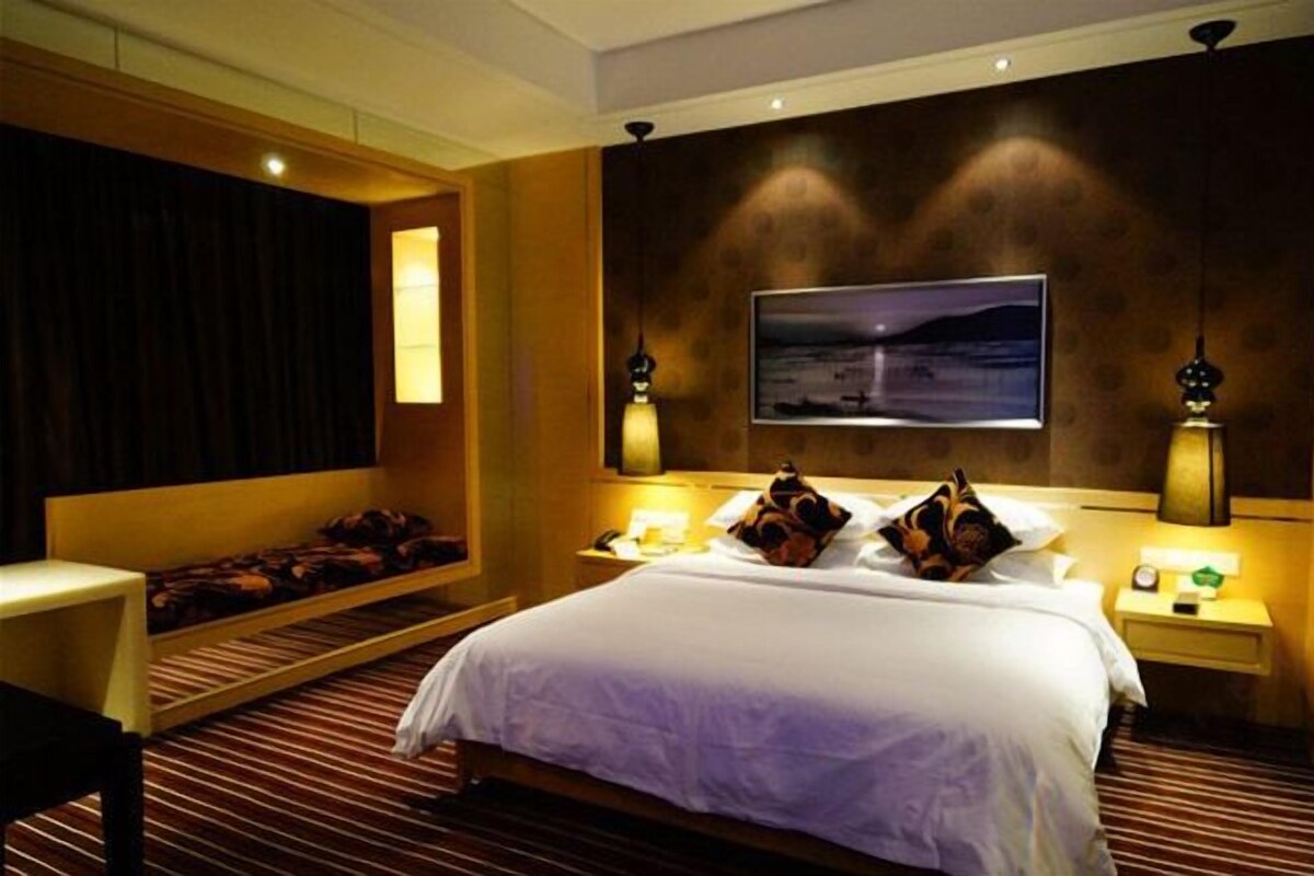 A hotel room with a large bed, a bedside table, a nightstand, a wall-mounted TV, and a window with curtains.