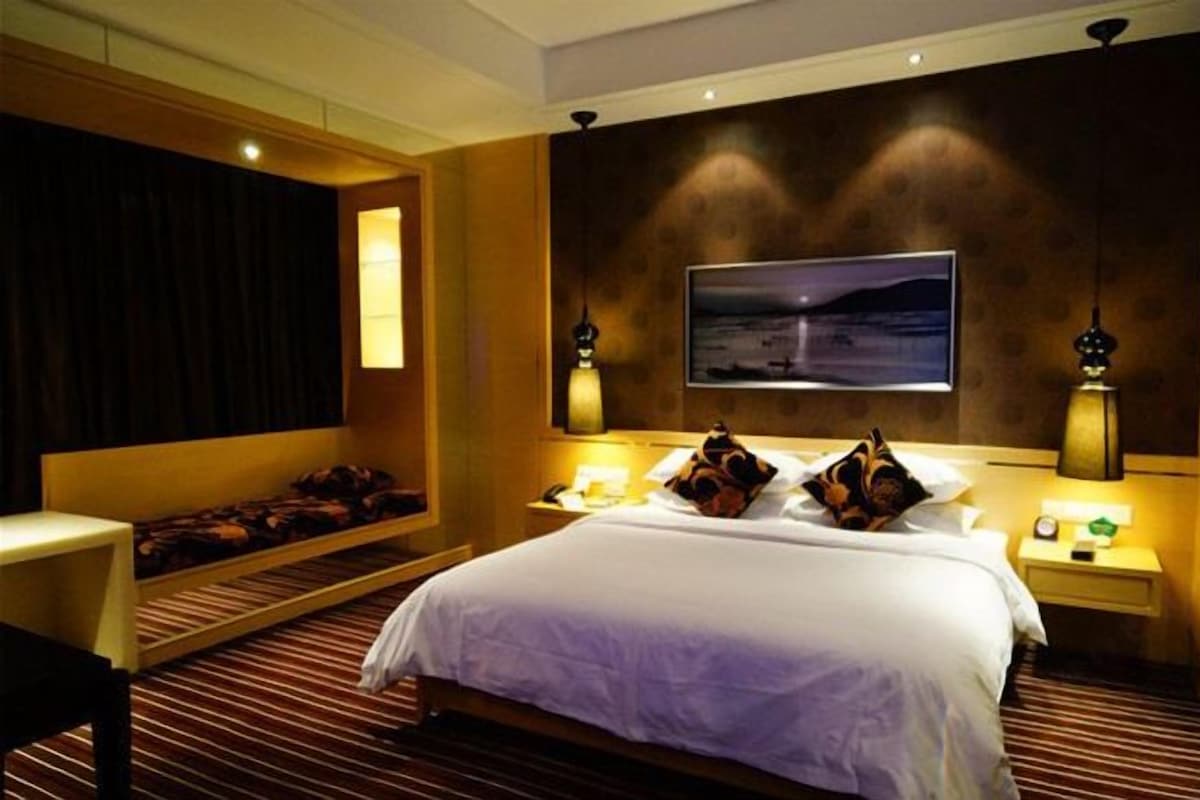 A hotel room with a large bed, a bedside table, a nightstand, a wall-mounted TV, and a window with curtains.