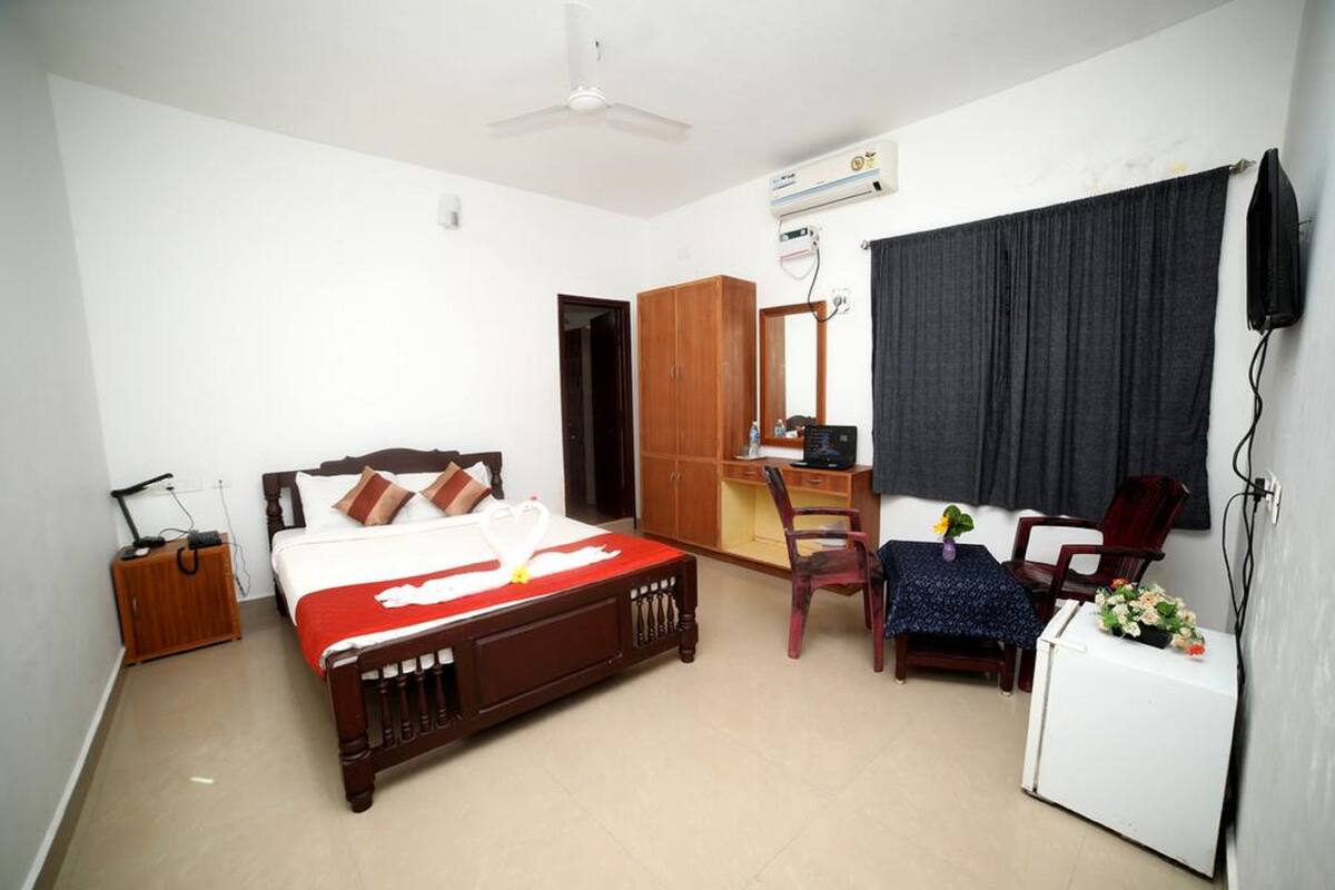 Quarto com cama de madeira, escrivaninha, cadeira, guarda-roupa, uma pequena mesa com flores e televisão.