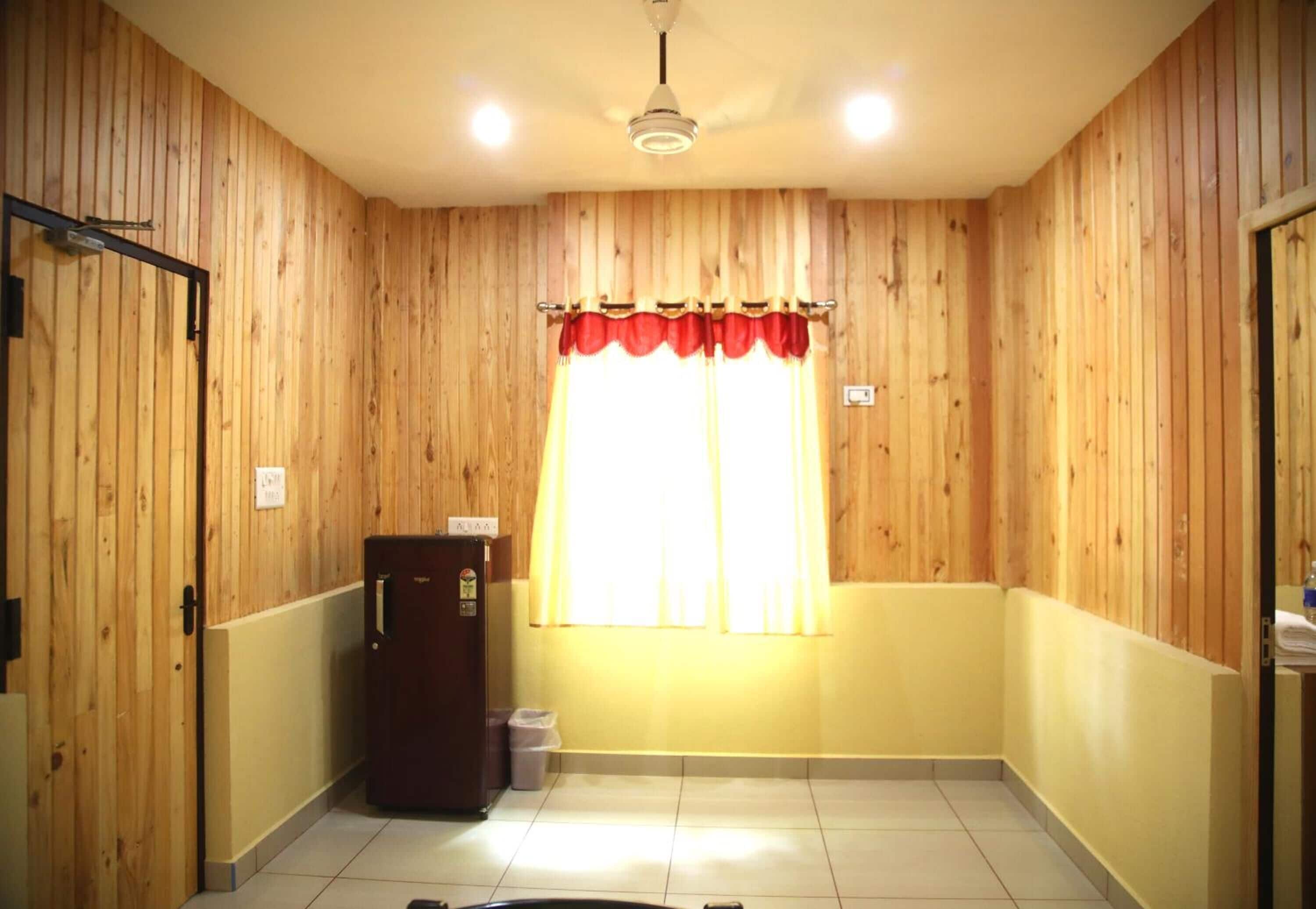 A room with wooden panel walls, a window with red curtains, a refrigerator, and a trash bin.