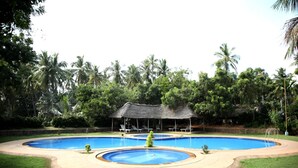 Une grande piscine extérieure, agrémentée d’un pavillon à toit de chaume et entourée d’une végétation luxuriante.