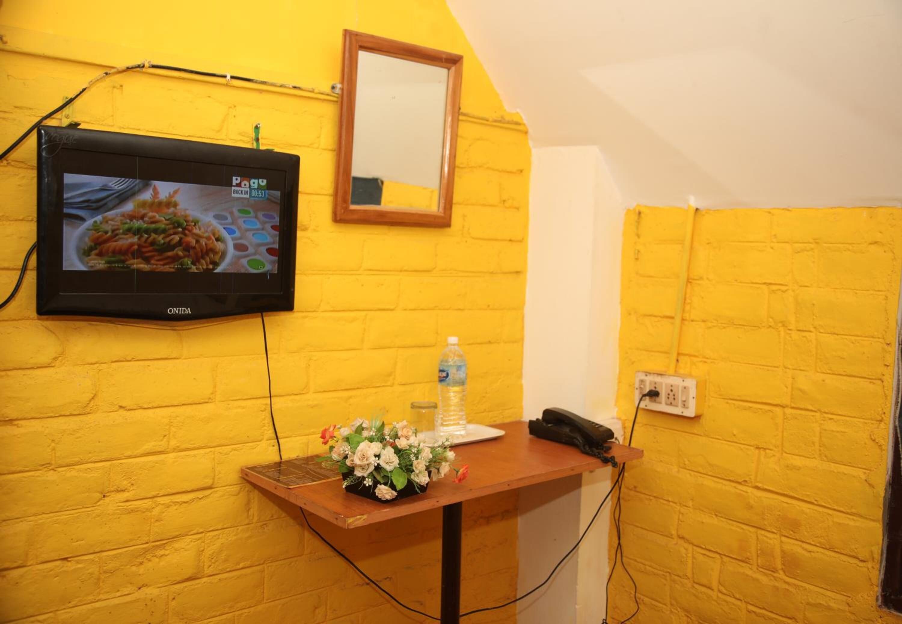 A room with a yellow brick wall, a mounted TV, a wooden desk with a water bottle, a phone, and a flower arrangement.