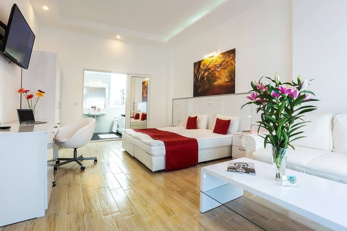 A modern hotel room with a bed, a desk with a laptop, a white sofa, and a large mirror.