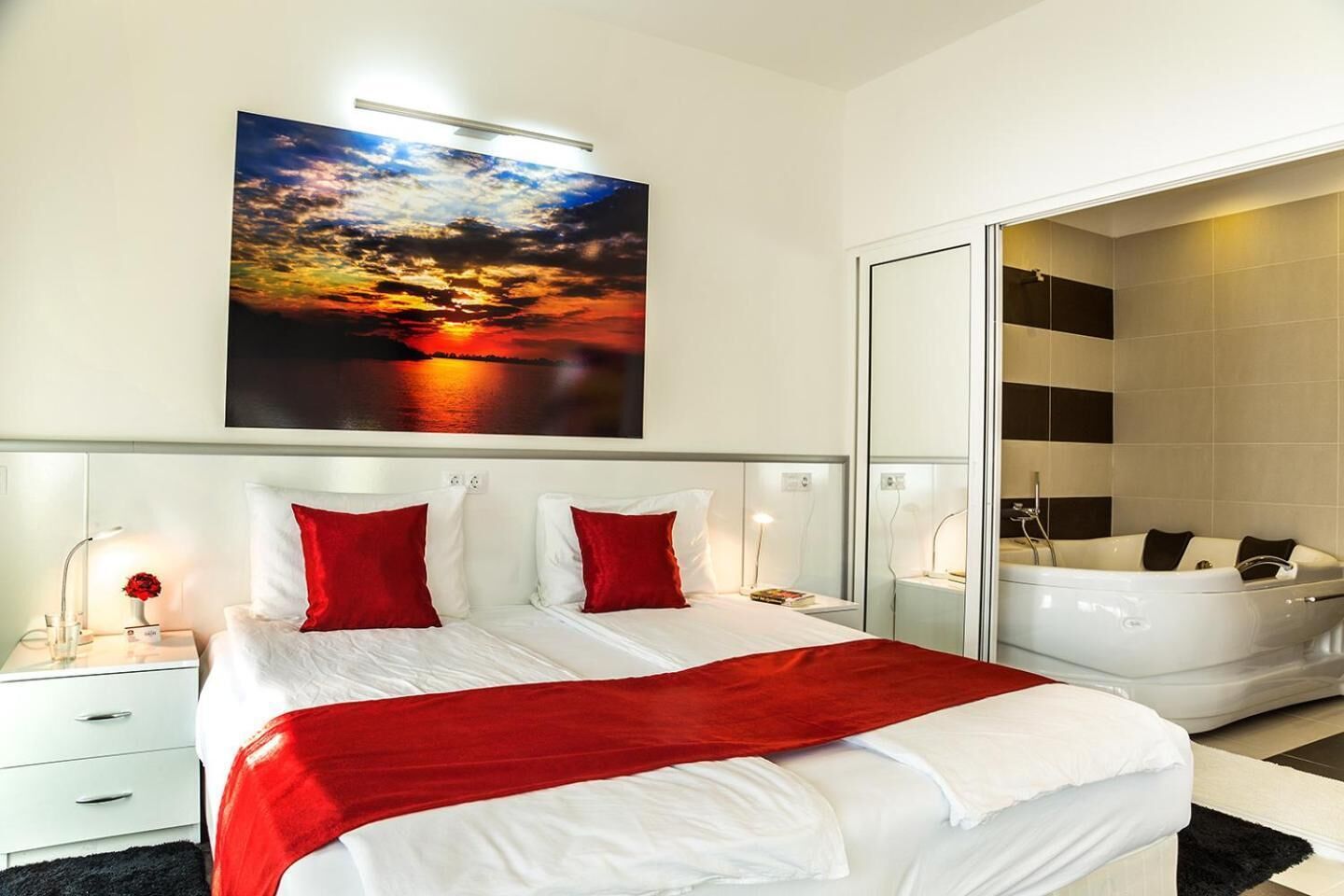 Una habitación de hotel moderna con una cama grande, almohadas rojas, una manta roja, un cuadro de un atardecer, un baño independiente con bañera y un espejo.