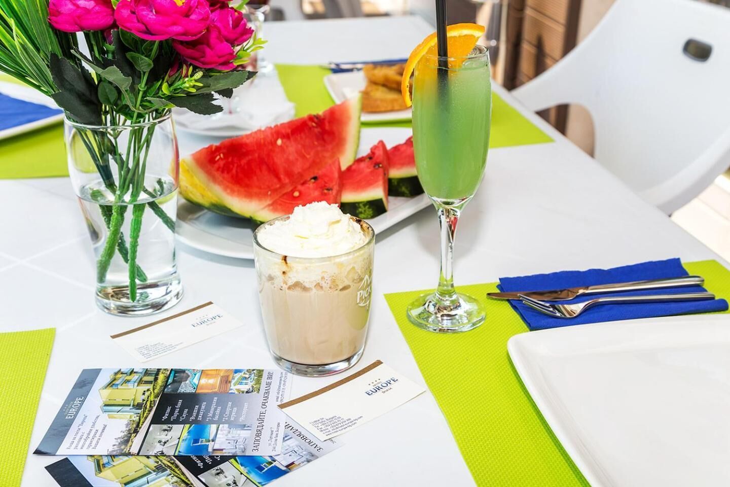 A table with a glass of iced tea, a glass of milkshake, a slice of watermelon, and a vase of flowers.
