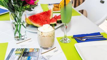 A table with a glass of iced tea, a glass of milkshake, a slice of watermelon, and a vase of flowers.