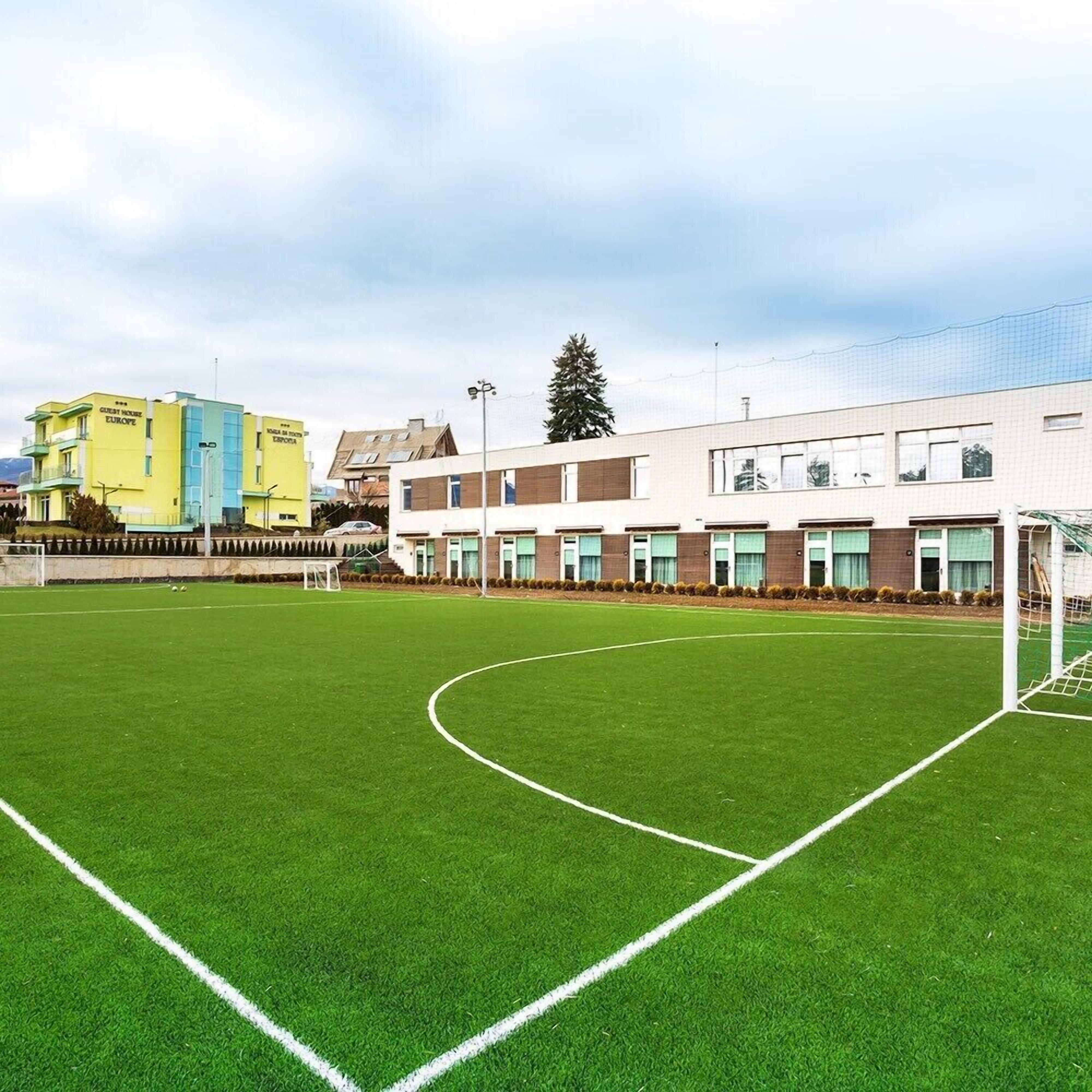 A soccer field with a goalpost, surrounded by buildings including a yellow hotel and a modern building with multiple windows.