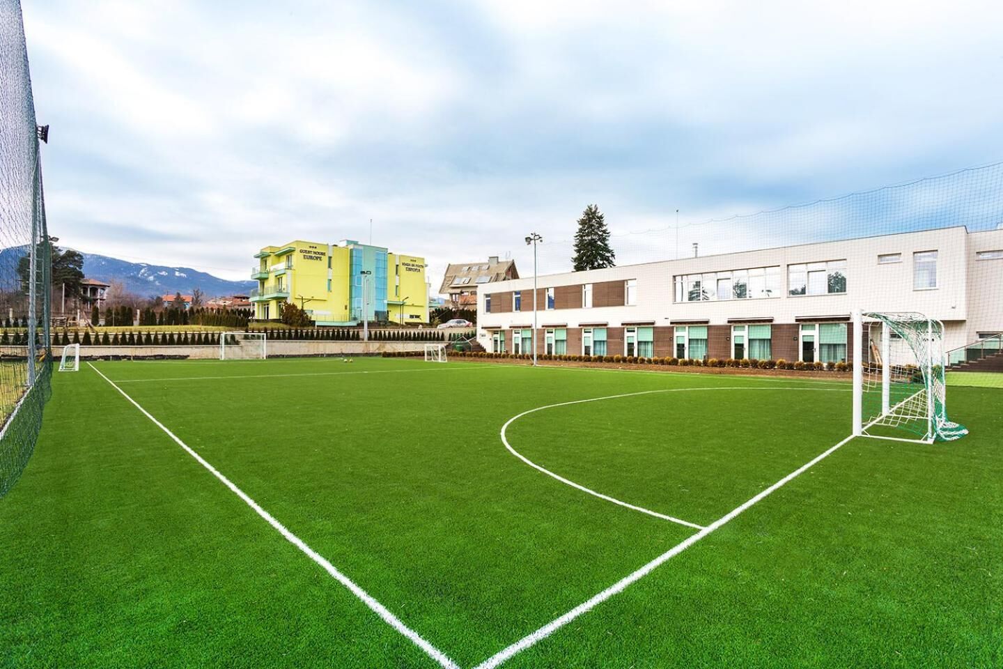 A soccer field with goalposts, surrounded by buildings with windows and a mountain in the background.