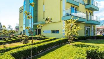 Un edificio de varios pisos con fachada amarilla, un letrero que dice "КВИЛА 3А РОСТИ ЕБРОПЛА", y un jardín bien cuidado con setos y árboles.