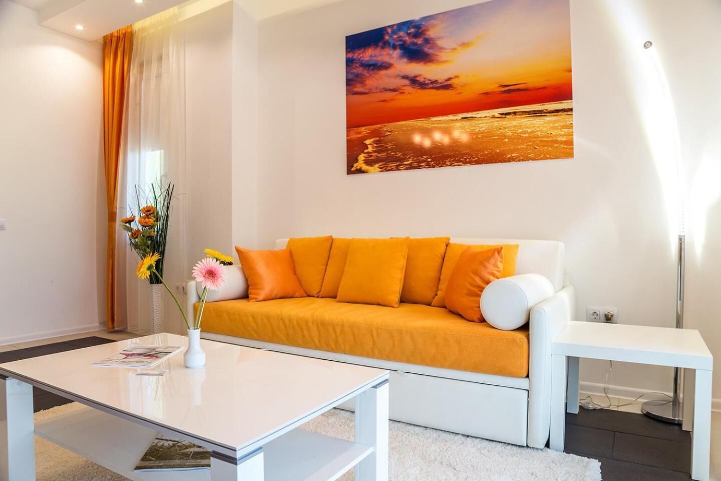 A modern living room with a white sofa, orange cushions, a glass coffee table, and a painting of a sunset over the ocean.