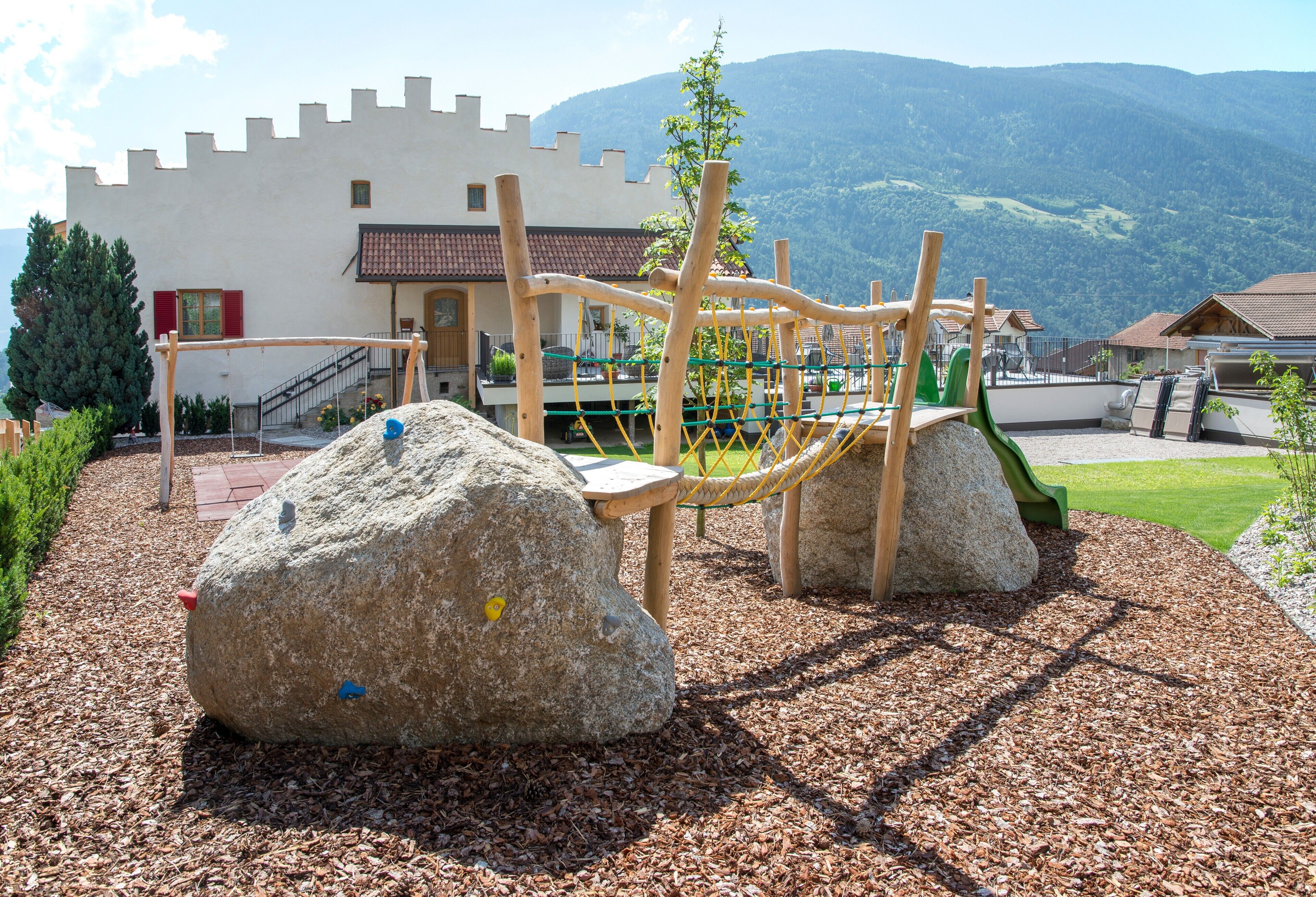 Um parquinho com estruturas de escalada e pedras grandes, com um edifício semelhante a um castelo e montanhas ao fundo.