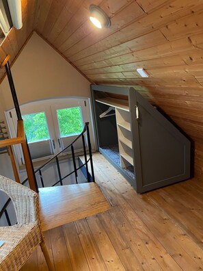A wooden interior with a staircase, a wicker chair, and a view of greenery outside.