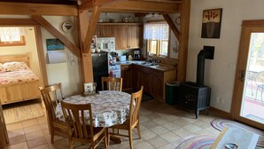 A cozy cabin interior with a wooden dining table, chairs, a kitchen area with a stove, and a bed with floral bedding. - Wooden guest cottage (East Glacier Park Village)