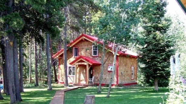 A wooden cabin with a red roof, surrounded by trees and a grassy yard. - Wooden guest cottage (East Glacier Park Village)