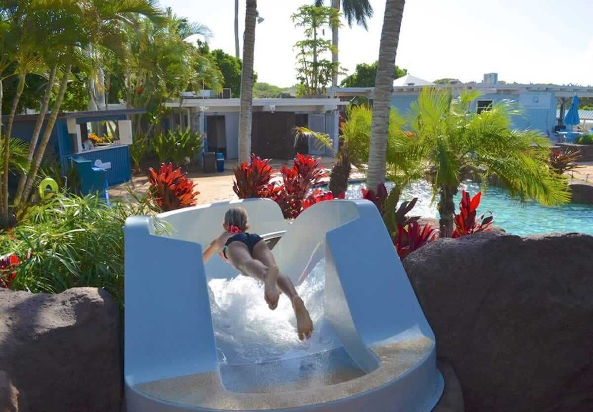Un tobogán de agua en una zona turística con palmeras y una piscina al fondo.