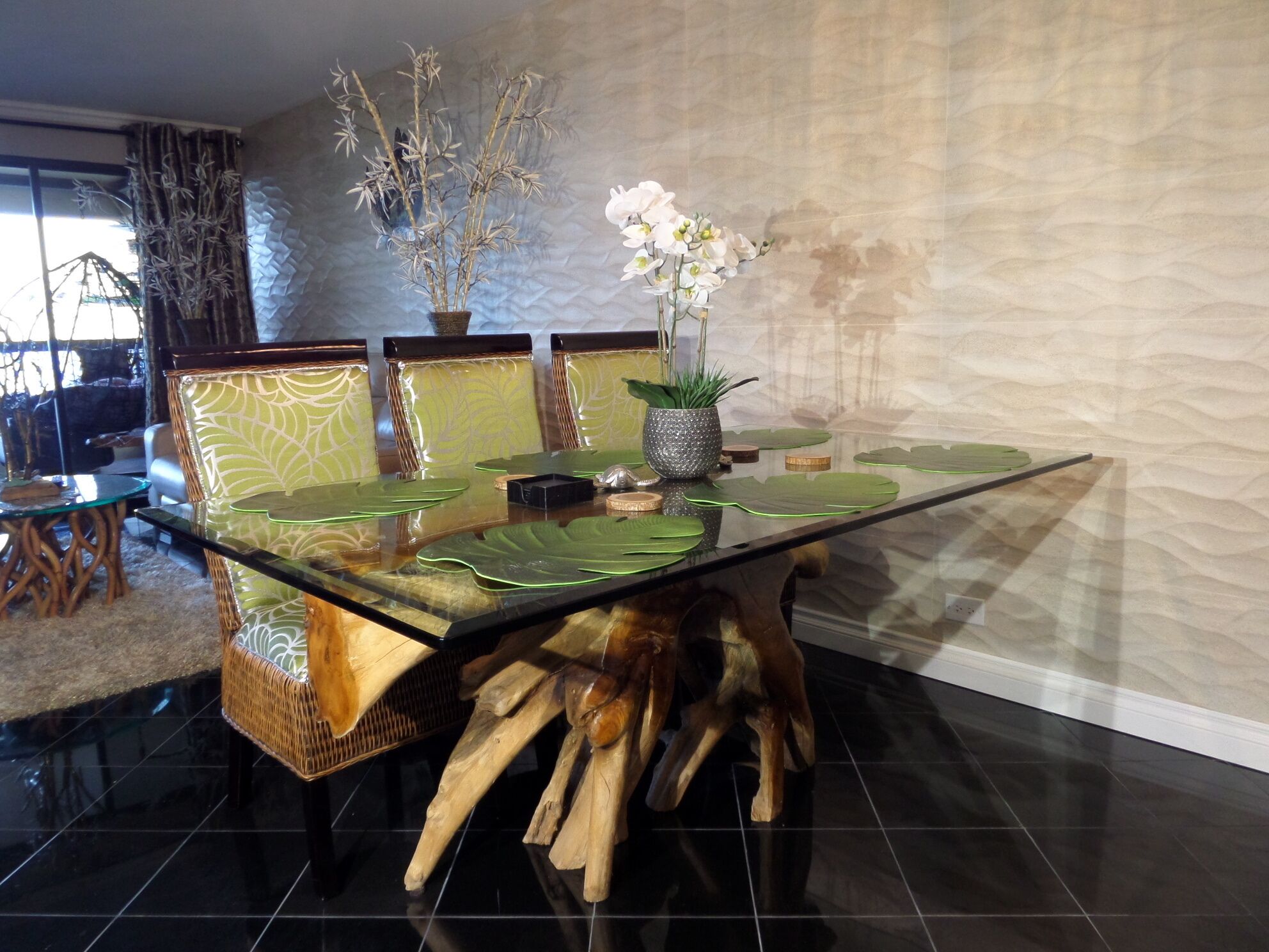A dining area with a glass table, green leaf placemats, and chairs with leaf-patterned upholstery.