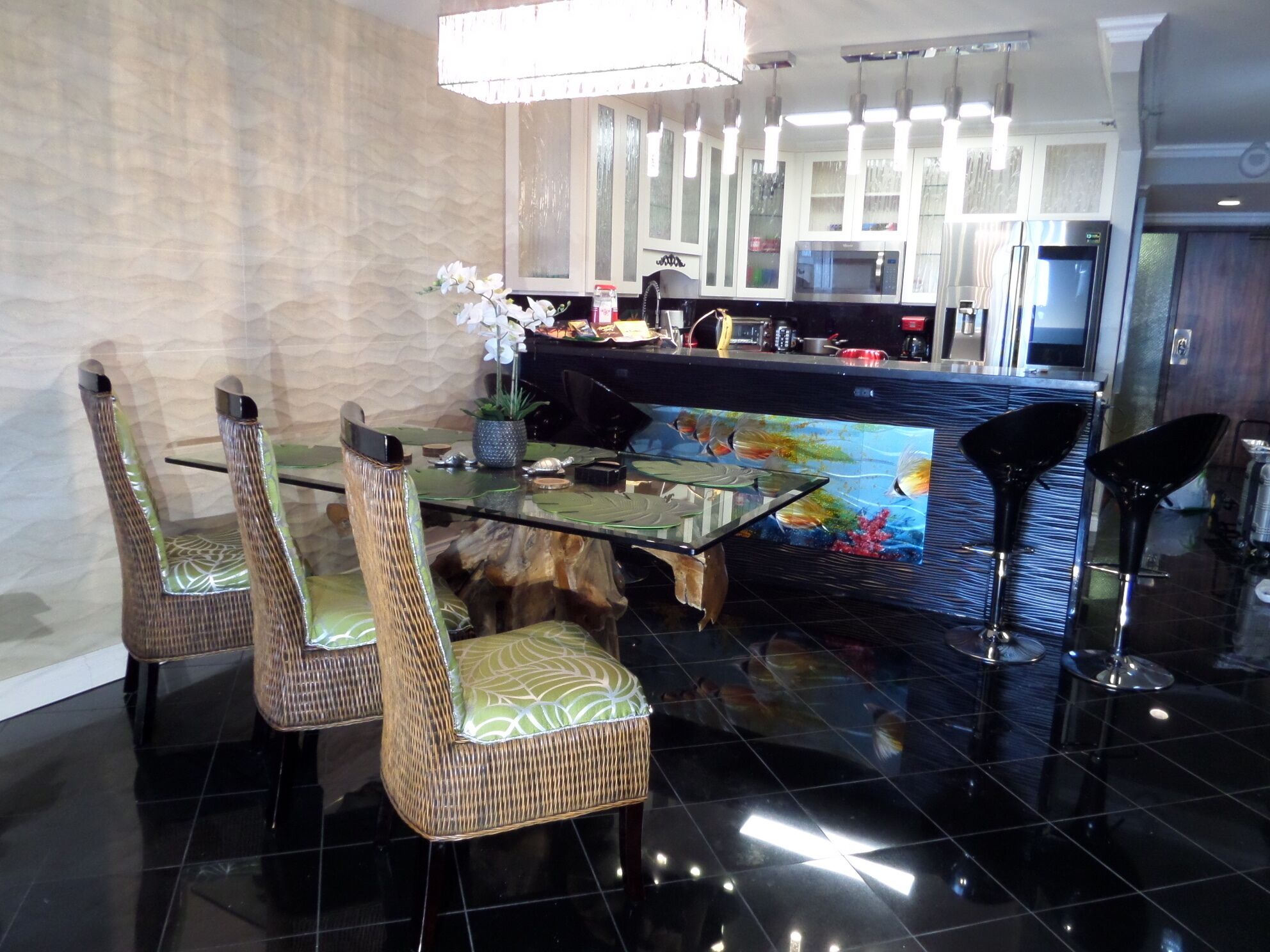 Una cocina moderna con una isla central, suelo de baldosas negras y un comedor con sillas de mimbre y una mesa de cristal.