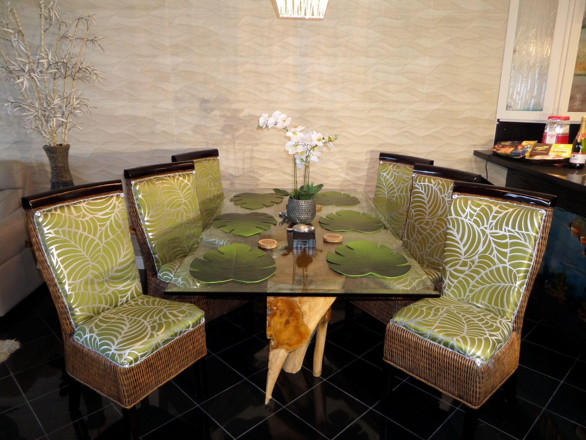 Une table à manger avec un plateau en verre, entourée de chaises en osier ornées de coussins à motifs verts.