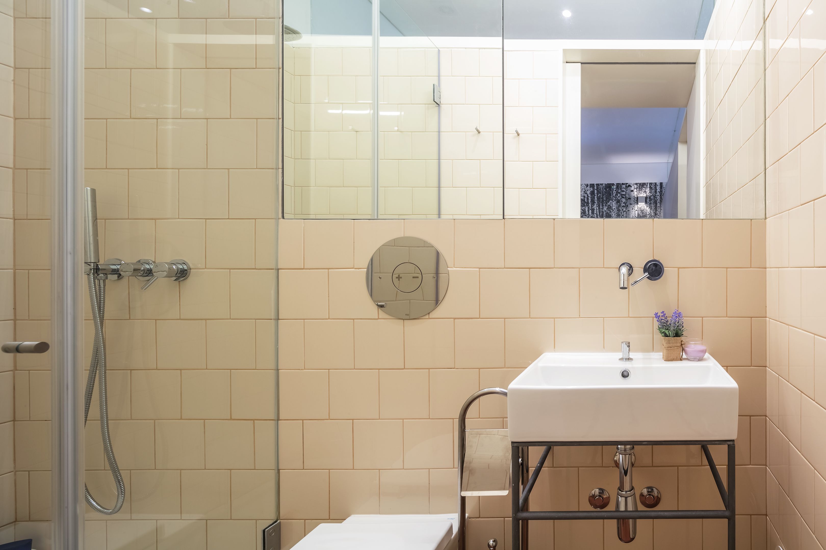 Ein Badezimmer mit Duschkabine aus Glas, einem weißen Waschbecken mit Spiegel darüber und beige gefliesten Wänden.