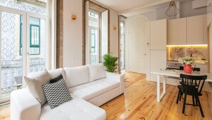 A modern living room with a white sofa, a dining table with a black chair, and a kitchen area with white cabinets and a marble backsplash. - Indulge Porto Flats (Porto)