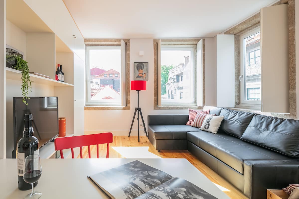 Een moderne woonkamer met een zwarte leren bank, een rode fauteuil, een eettafel met een fles wijn en een glas, en een groot raam met uitzicht op gebouwen.