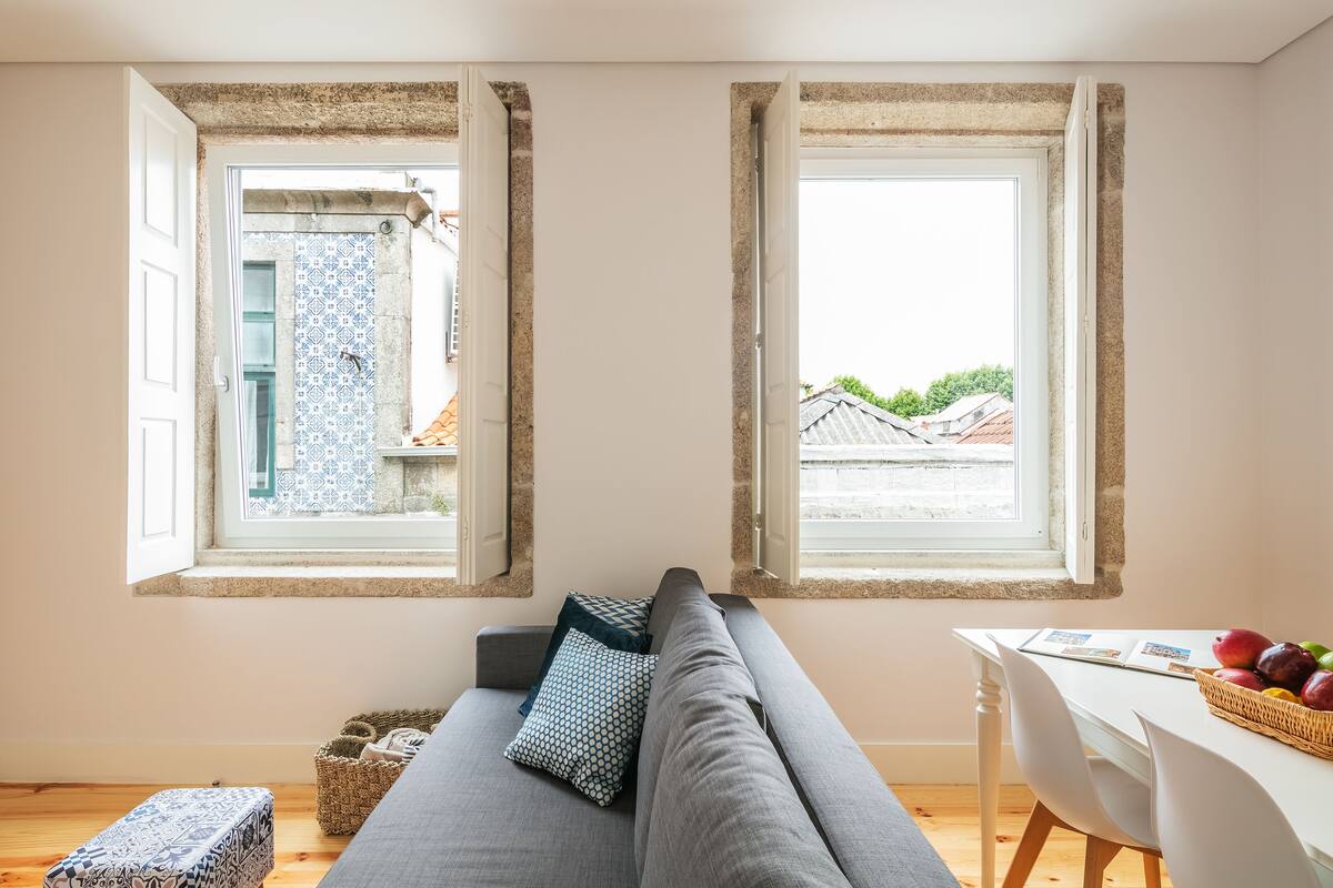 Een gezellige kamer met een grijze bank, twee ramen met behang, een witte tafel met een mand fruit, en een klein bankje.